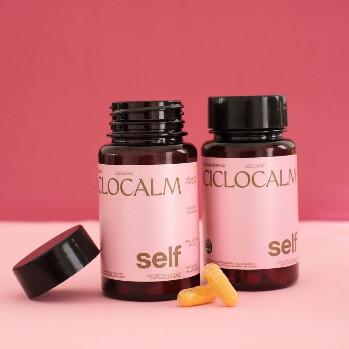 2 ciclocalms (suplemento para el dolor menstrual y ciclo femenino) sobre fondo rosa. Uno está abierto y el otro cerrado. Hay dos capsulas de ciclocalm en el frente.