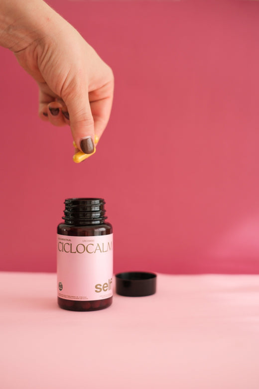 Ciclocalm (suplemento a base de magnesio, curcuma, jengibre y zinc, en capsulas, para el dolor menstrual) abierto sobre fondo plano. Hay una mano sosteniendo dos capsulas encima del producto. 