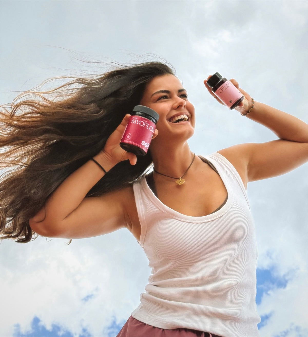 Mujer joven sosteniendo myoflow en una mano y ciclocalm en la otra. Foto tomada desde abajo, con fondo de cielo. Mujer en remera sin mangas con sonrisa y pelos al viento. Utiliza collar de corazon. 