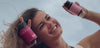 mujer sosteniendo un myoflow y un ciclocalm, sonriendo sobre fondo de nubes. Luego, myoflow en un vaso curvo de vidrio sobre fondo rosa plano . Luego, cclocalm y myo flow sobre fondo planorosa. Mujer joven con productos.  Myoflow y ciclocalm sobre plantas y flores rosas. 